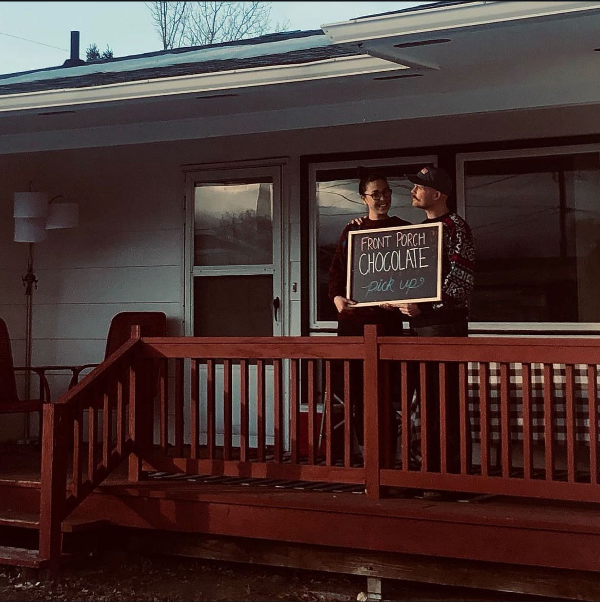 Two people on a porch holding a sign that says 'Front Porch Chocolate', with a house in the background.